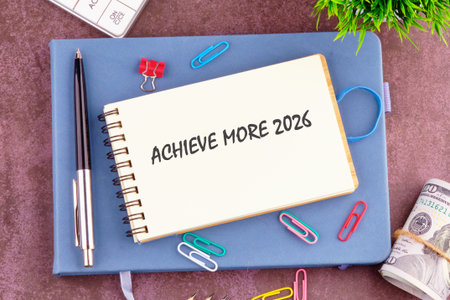 A notebook displaying the words Achieve More 2026 rests atop a blue journal. A pen, paperclips, money, and a calculator surround the notebook, suggesting planning and financial goals.の写真素材