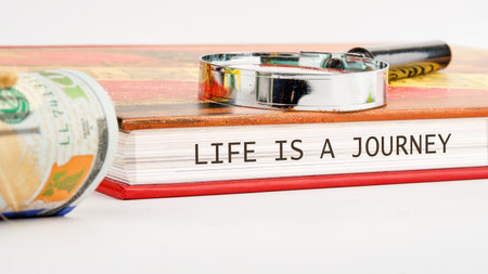 A roll of United States currency rests beside a book titled LIFE IS A JOURNEY. A magnifying glass rests on top of the book, symbolizing exploration.の写真素材