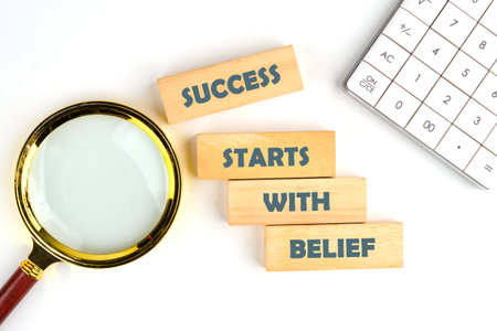 A magnifying glass hovers over wooden blocks spelling out the phrase success starts with belief...