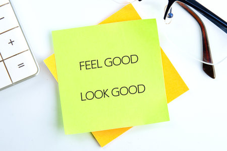 A vibrant sticky note with the message feel good look good sits on a wooden desk. Nearby, a calculator and glasses are placed, creating a motivational and organized workspace atmosphereの写真素材