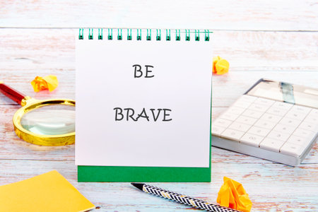A notepad featuring the words Be Brave rests on a wooden desk surrounded by crumpled paper, a pen, a magnifying glass, and a calculator, symbolizing motivation and creativityの写真素材