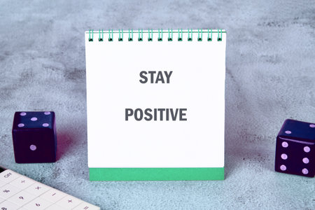 A small flip calendar displays the words stay positive on a soft, gray surface. Two colorful dice sit nearby, adding a playful touch to the uplifting message. Perfect for daily inspirationの写真素材