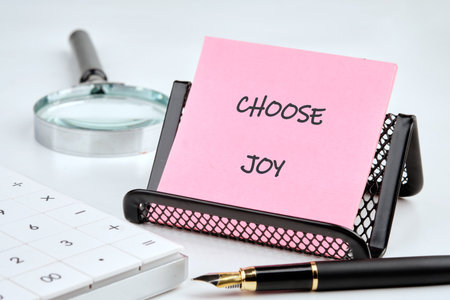 A bright pink note with the words Choose Joy rests in a black holder on a desk. Nearby, a magnifying glass and a pen are prepared for a day of work and creativity.の写真素材
