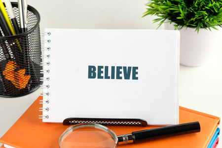 A notepad displaying the word BELIEVE inspires confidence and positivity. The scene features a magnifying glass, stationery, and a potted plant, creating a warm atmosphere for creativity.の写真素材