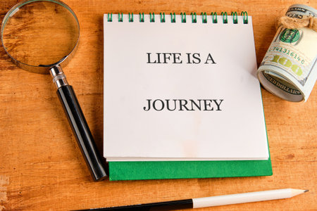 A notebook reveals the phrase life is a journey, resting on a wooden table alongside a magnifying...