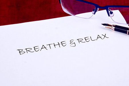 This shows a written reminder to Breathe and Relax on white paper. Blue framed glasses and a fountain pen are on the paper, set against a deep red background.の写真素材