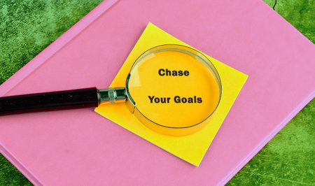 A magnifying glass highlights the phrase Chase Your Goals written on a vibrant yellow sticky note,...