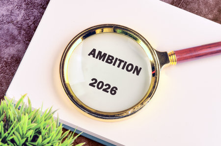 A magnifying glass focuses on the words AMBITION and 2026, sitting on a white book, next to a small...