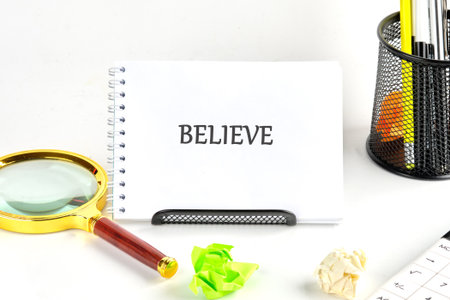 A notebook displaying the word BELIEVE sits on a white surface, accompanied by a golden magnifying...
