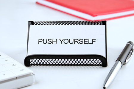 A motivational message is displayed on a white card in a black holder next to a red notebook, a calculator, and a pen. The card reads PUSH YOURSELF.の写真素材
