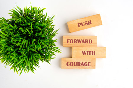 On a clean white surface, wooden blocks are stacked saying, PUSH FORWARD WITH COURAGE, along with a...