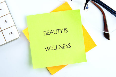 On a clean white desk, yellow and lime green sticky notes display the words Beauty Is Wellness, alongside eyeglasses and a calculator. A reminder for health and self-care.の写真素材