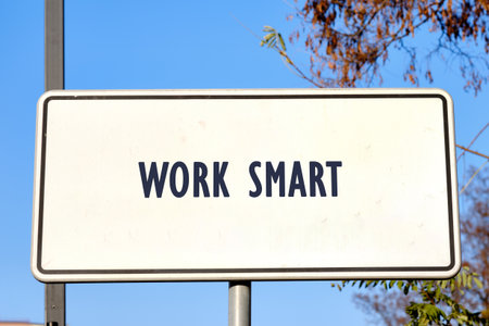 A white sign displays the words Work Smart in dark blue lettering. The sign is mounted on a pole, set against a radiant blue sky with trees in the background.の写真素材