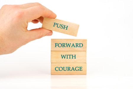 A hand places a wooden block labeled PUSH atop a tower of blocks reading FORWARD WITH COURAGE....
