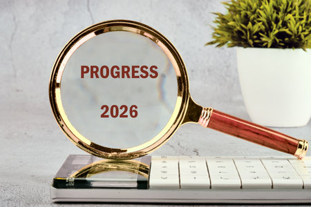 A magnifying glass is poised over a calculator, highlighting the words PROGRESS and 2026. A small potted plant adds a touch of nature to the compositionの写真素材
