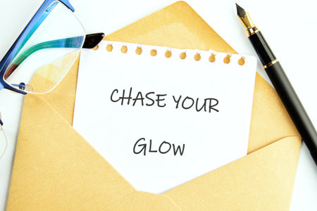 A paper with text CHASE YOUR GLOW sits in an open gold envelope near blue glasses and a black...