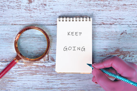 On a rustic wooden surface, a hand holds a pencil next to a notepad displaying the phrase KEEP...
