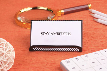A white card displaying Stay Ambitious is prominently placed on a vibrant orange textured desktop.