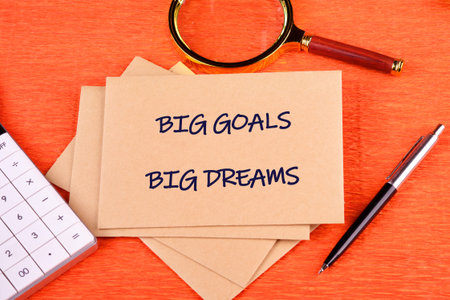 A stack of light brown envelopes shows BIG GOALS BIG DREAMS on an orange surface.