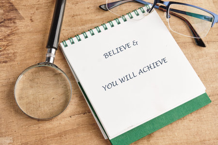 Motivational quote Believe and You Will Achieve written on a white notepad on a wooden desk. A...