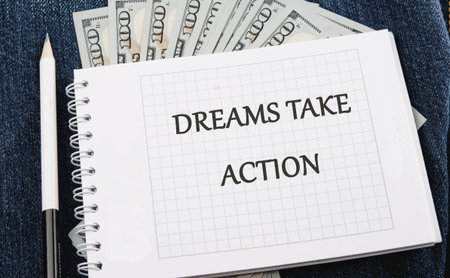 A white spiral notebook shows DREAMS TAKE ACTION on its page. One hundred United States dollar bills...