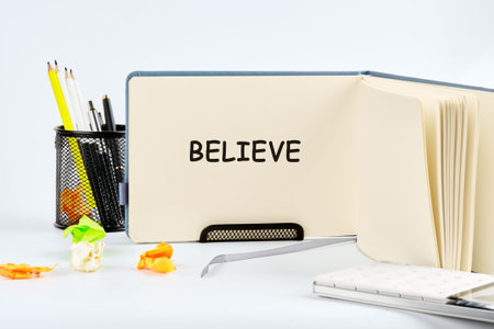 BELIEVE in an open notebook on a desk with pens, pencils, crumpled paper. Motivates work study.