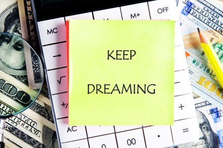 Bright yellow sticky note with the words Keep Dreaming placed on a calculator surrounded by cash and...