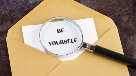 A gleaming magnifying glass rests on a white sheet, revealing the encouraging words BE YOURSELF, nestled within a golden envelope upon a textured dark surface.の写真素材