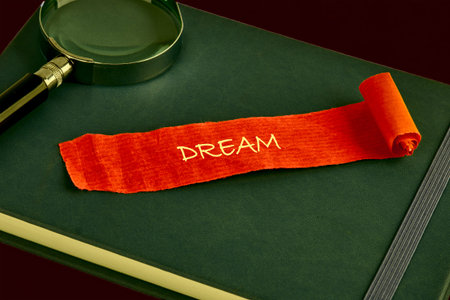 A green leather journal rests on a dark surface, holding a bright orange fabric strip embroidered with the word DREAM.の写真素材