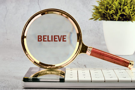 Gold wood magnifier focuses red Believe on white calculator on gray surface, near green plant