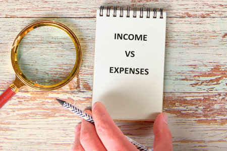 A focused hand, holding a stylish pencil, hovers over a notepad explicitly marking INCOME VS...