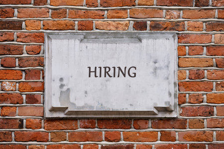 A hiring sign is showcased on a brick wall. The sign looks weathered and is placed in a busy commercial area. People pass by during the day.