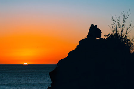 Boy and girl on a rock watching the sunset, silhouettes of couple watching the sunsetの写真素材