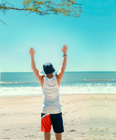 man with hat from back on the beach, young man from back raising his hands on the beach.