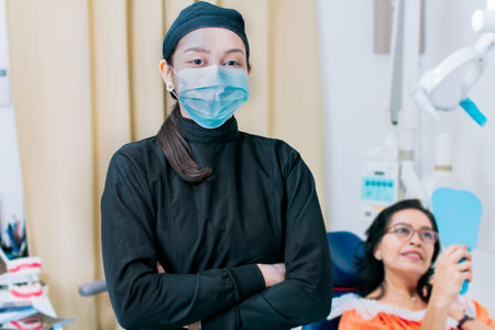 Dental doctor standing in clinic wearing mask, portrait of dentist crossing arms with patient in background, Modern Dental clinic. Dental procedures. portrait of a dentist woman with crossed armsの写真素材