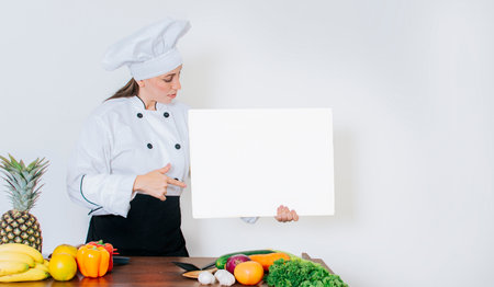 Chef woman with vegetables at table holding blank menu. Girl chef in the kitchen showing a blank board, Beautiful woman chef with table of vegetables holding a blank boardの写真素材