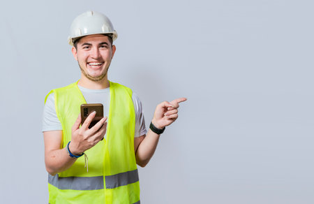 Happy Civil engineer holding cellphone and pointing a promo. Happy engineer holding phone and pointing to the side isolated. Young construction engineer holding phone and pointing at promoの写真素材