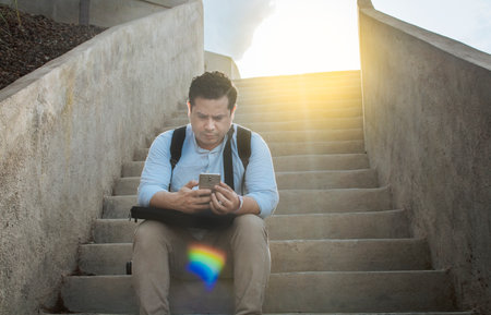 Handsome man sitting on stairs using smart phone, Front view of guy sitting on stairs using his cell phone outdoors, Close up of young man sitting on stairs using mobile phone outdoorsの写真素材
