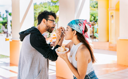 A couple eating shaved ice in a square, Young couple eating shaved ice together with arms intertwined, Side view of young couple with arms linked eating ice cream in a city squareの写真素材