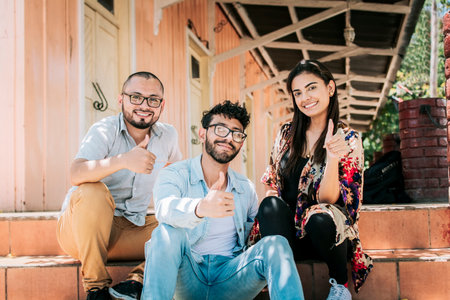 Group of happy friends sitting on the stairs giving thumbs up. Portrait of happy teenagers sitting outside giving thumbs up, Close up of happy sitting students giving thumbs upの写真素材