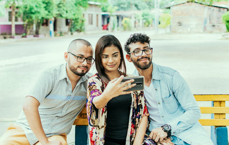 Three friends taking a selfie on a park bench, Three smiling friends sitting on a bench taking a selfie. Front view of three happy friends taking a selfie while sitting on a benchの写真素材