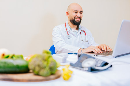 Smiling nutritionist with laptop at desk. Nutritionist doctor using laptop at workplace. Bearded nutritionist doctor working on laptop at desk.の写真素材