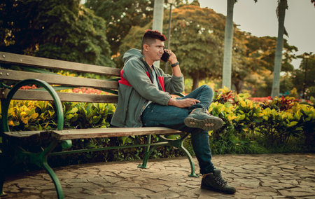 Man sitting on a bench calling on the phone, young man sitting on a bench with cell phone, close up of a young man calling on the phone sitting on a benchの写真素材