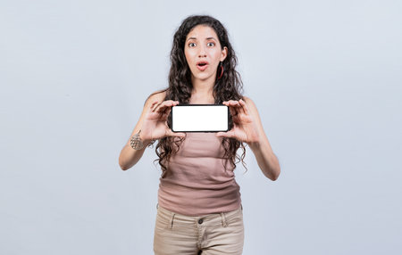 Surprised girl showing blank screen of smart phone. Latin girl showing horizontal screen of cell...