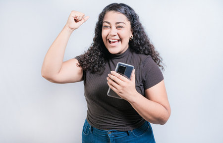 Winning happy young woman holding cell phone. Happy latin girl celebrating with phone isolated. Happy people holding smartphone and celebratingの写真素材