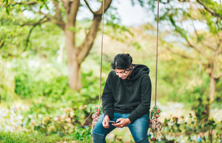 Smiling guy sitting on a swing with smart phone. Young man sitting on a swing using cell phone in natureの写真素材