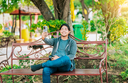 Relaxed guy taking a selfie sitting on a bench at sunset. Smiling man sitting on a bench taking a selfie in Nagarote Parkの写真素材