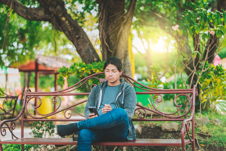 Handsome man sitting on a bench checking cell phone at sunset. Young man sitting on a park bench texting on cell phoneの写真素材