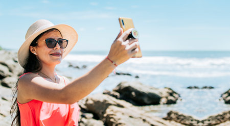 Girl in hat on the beach taking a selfie. Young woman on vacation taking photos on the beach, Smiling girl in hat taking photos on the beach. Beach vacation conceptの写真素材