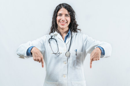 Smiling female doctor pointing down at a space isolated. Young female doctor showing something downの写真素材
